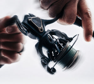 A close-up action shot of a black spinning fishing reel being held by two hands against a bright white background with motion blur.