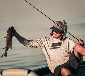 A man in a boat on the water holds up a small fish while wearing a Abu Garcia branded long-sleeve performance shirt, dark shorts, and a cap.