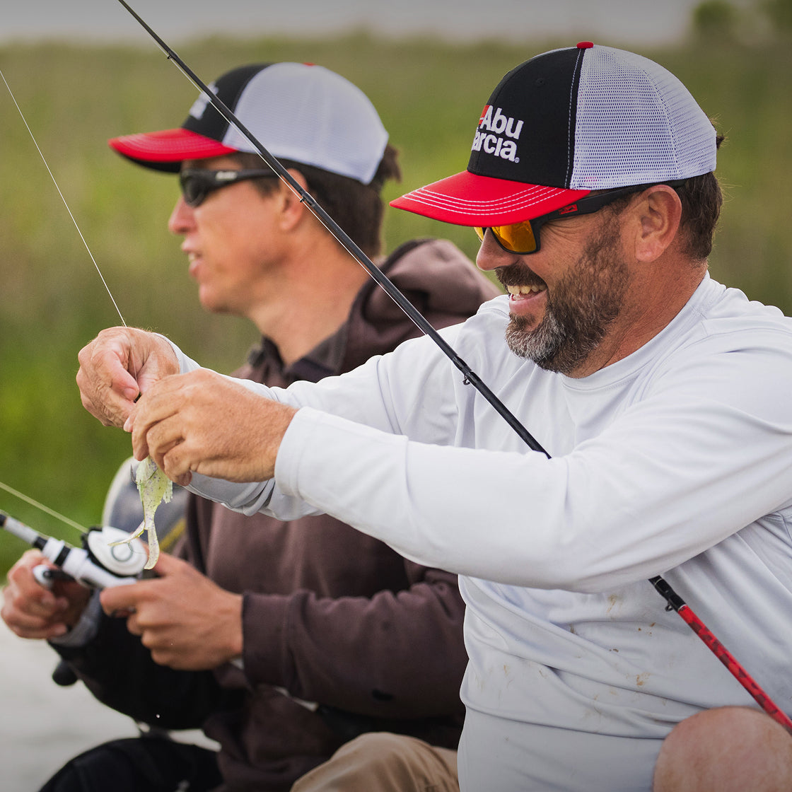 Two men on a boat wearing Abu Garcia branded apparel with one holding a fishing rod and the other holding a fish, surrounded by greenery.