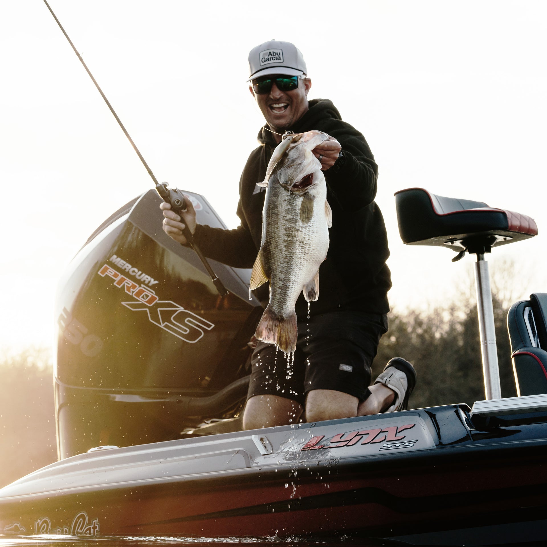 Man on a boat holding a bass in one hand and a low profile baitcast combo in the other, sporting an Abu Garcia branded hat and sunglasses. 