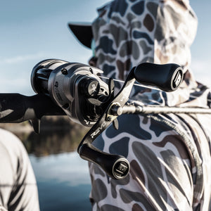  Revo® VoltiQ™ Low Profile Reel held by a person wearing a camouflage hoodie with a blurred natural background