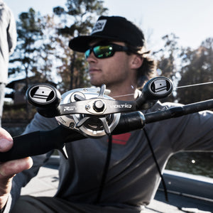 Close-up of a man wearing an Abu Garcia Branded hat & shirts wearing sunglasses holding the Revo® VoltiQ™ Low Profile Reel with a blurred background
