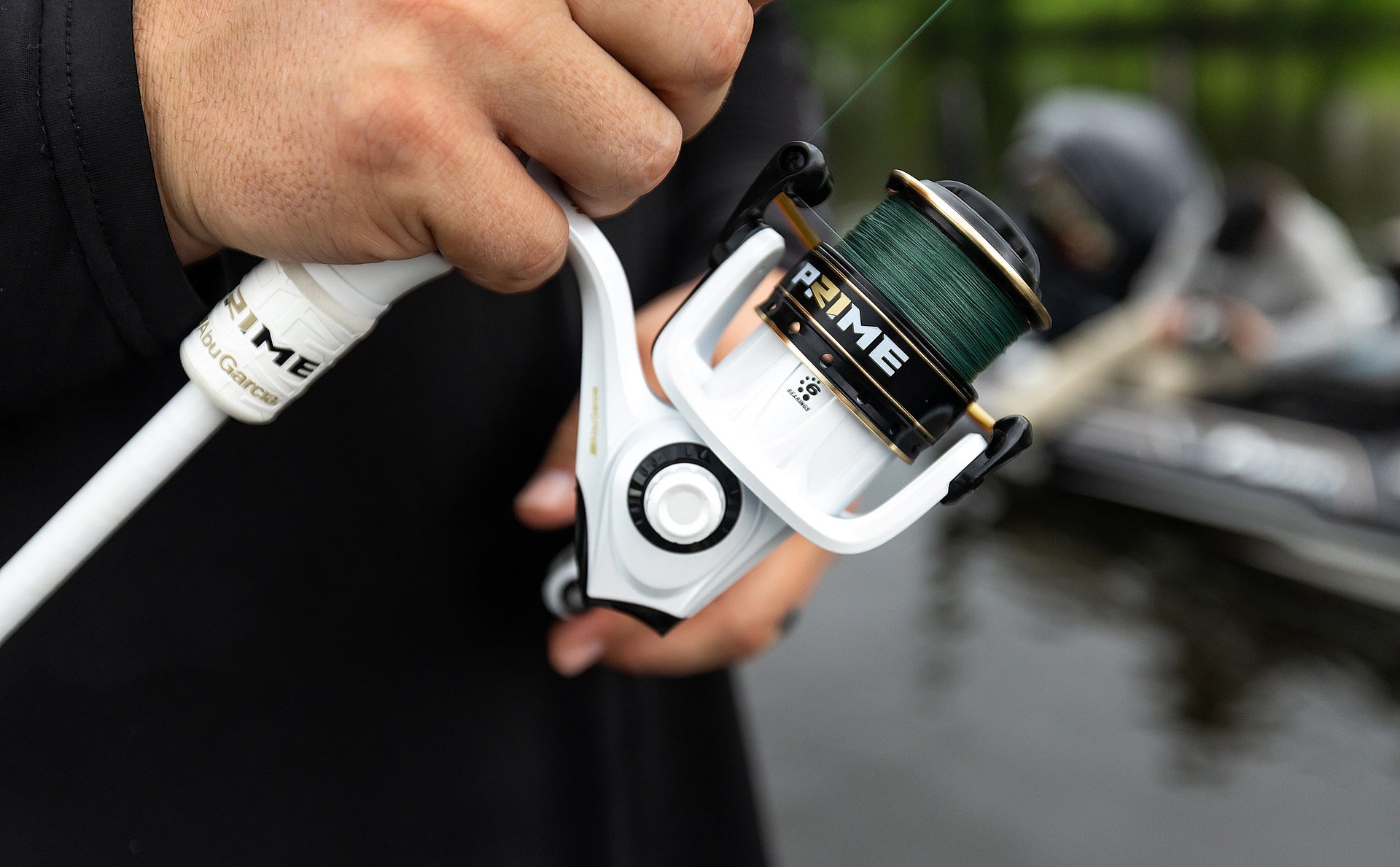 Fishing reel with Prime branding held by a person, blurred background of water and boat.