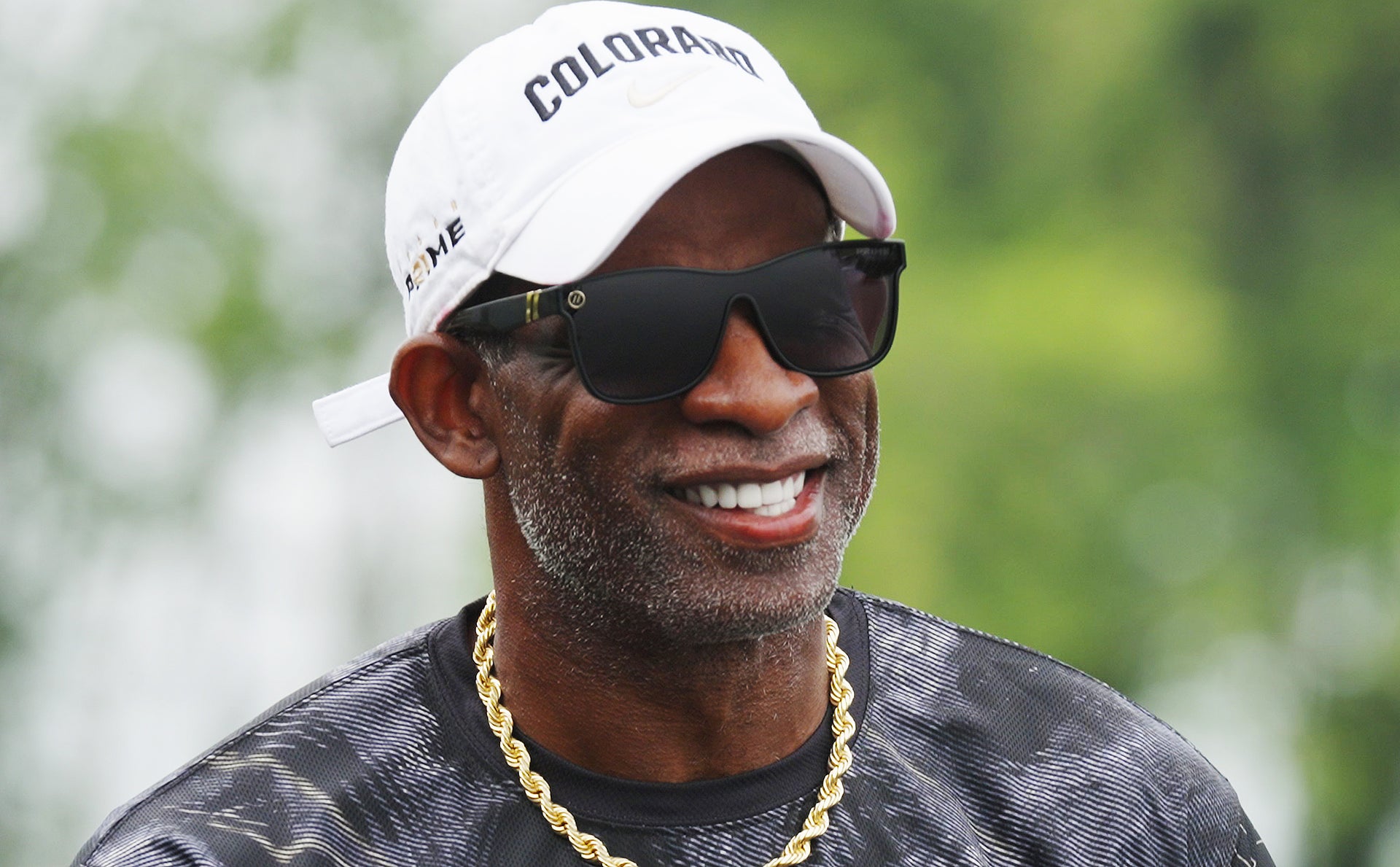 Man wearing a white cap with 'Colorado' text, sunglasses, and a gold chain necklace against a blurred green background.