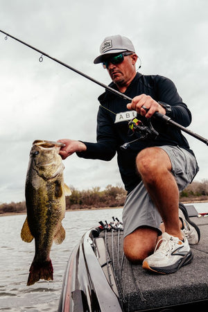 A man kneeling in a boat on the water holds up a large bass fish while holding a Abu Garcia Veritas Tournament Solid Tip fishing rod. He is wearing a gray cap, sunglasses, a black long-sleeve shirt, gray shorts, and white sneakers. 