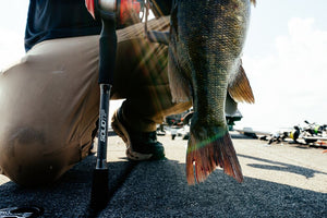 A close-up action shot of a person kneeling on a boat deck holding a freshly caught largemouth bass, with a Abu Garcia Veritas Tournament Solid Top Rod standing nearby.